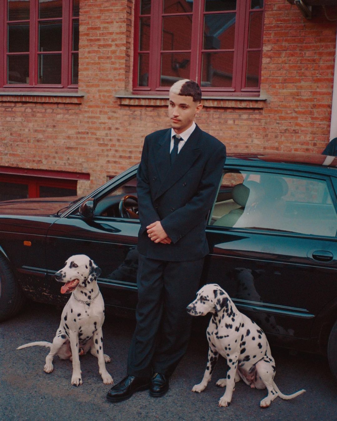 Young adult male in suit poses between two dalmatian dogs near a black car and a brick building with red window frames in daytime.