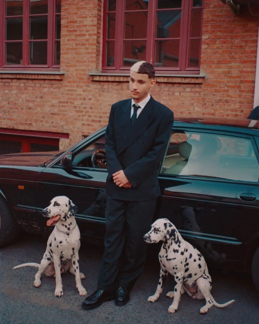 Young adult male in suit poses between two dalmatian dogs near a black car and a brick building with red window frames in daytime.