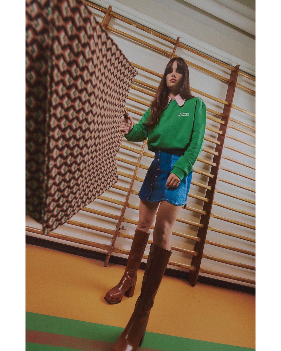 A young woman poses in a bright green sweater, blue denim skirt, and brown knee-high boots in what appears to be a gymnasium.