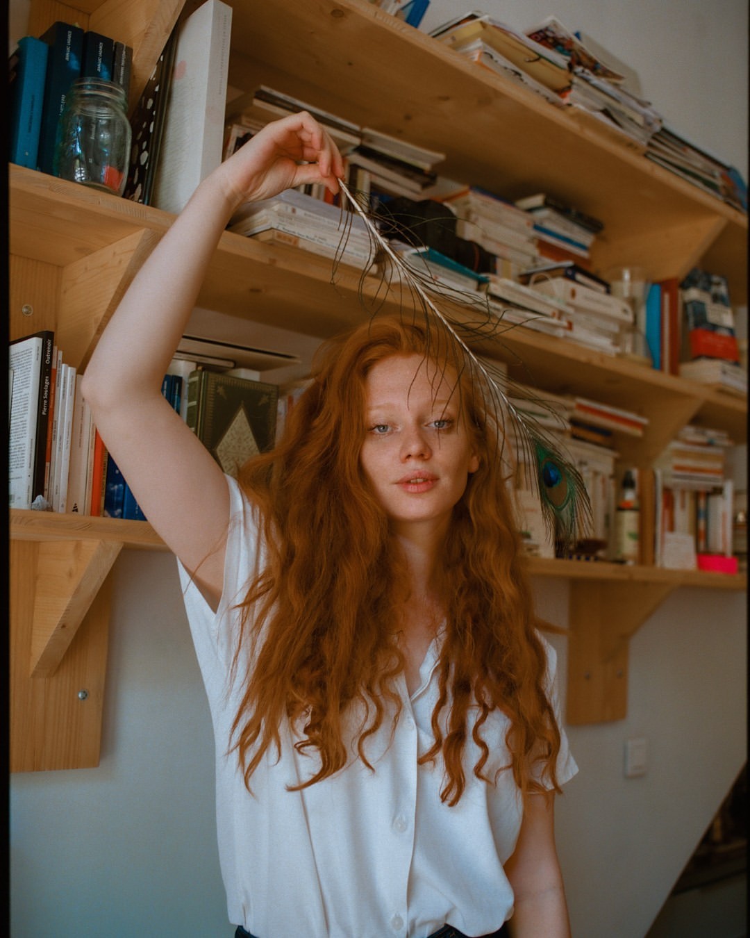 A young woman with long red hair poses indoors while holding a peacock feather. The backdrop features a bookcase filled with books.