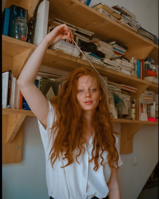 A young woman with long red hair poses indoors while holding a peacock feather. The backdrop features a bookcase filled with books.