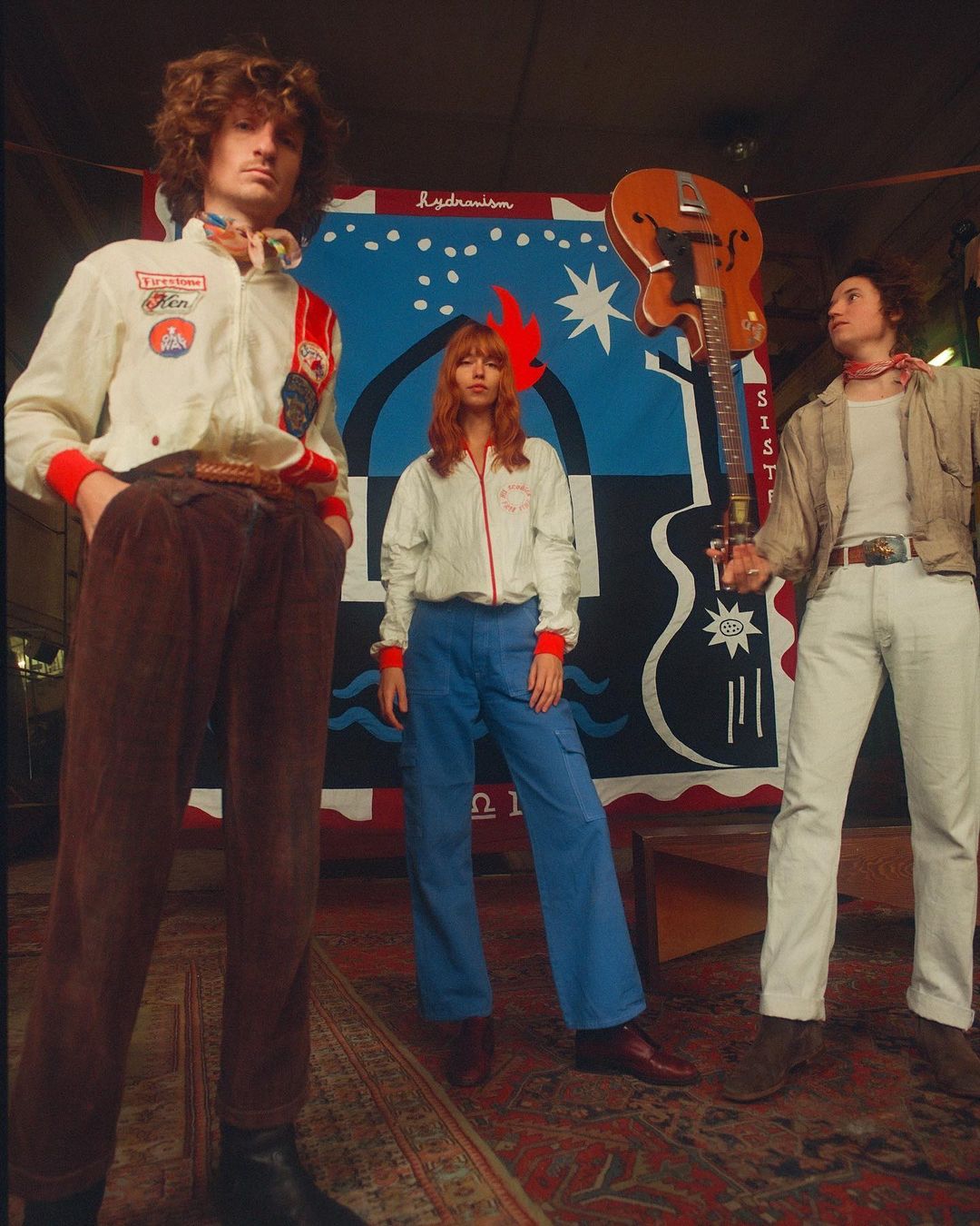 Three young adult musicians in vintage clothing pose in front of a vibrant, hand-painted backdrop with a vintage guitar. 