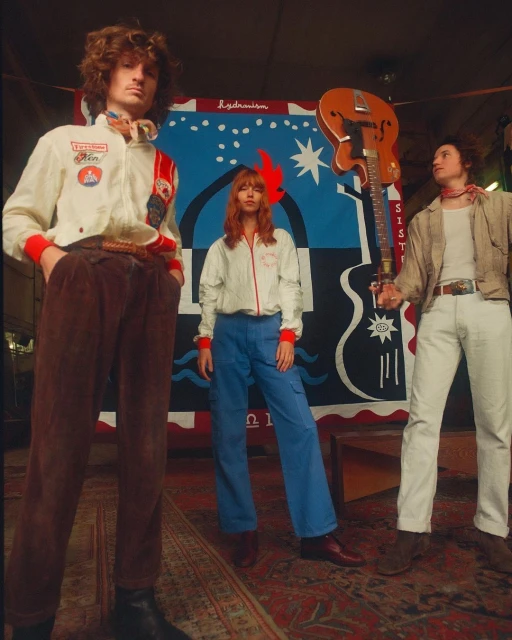 Three young adult musicians in vintage clothing pose in front of a vibrant, hand-painted backdrop with a vintage guitar. 