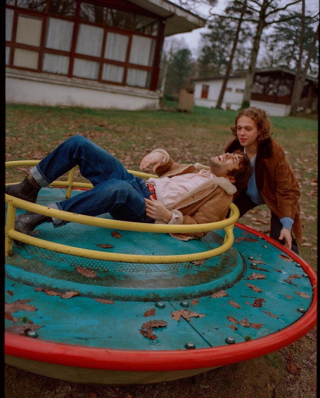Two adults on a vintage merry-go-round on a grassy field, one pushing and one laying back laughing on a cloudy day.