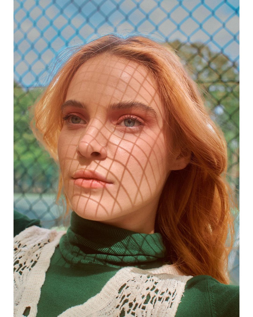 Portrait of a woman with light red hair in a green top with fence shadow on her face, set against a blue chain-link fence.