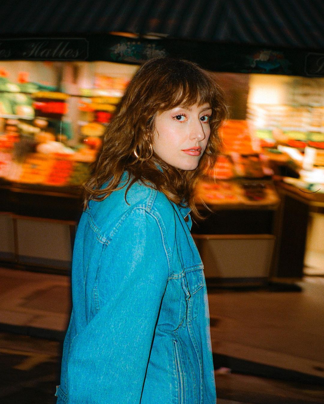 Portrait of a young adult woman with long wavy brown hair and a denim jacket stands in front of a blurred background of a store.