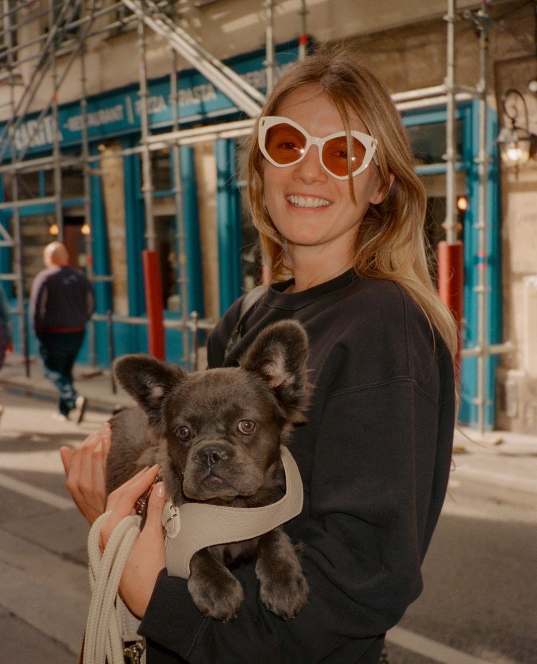 A woman in sunglasses smiles while holding a French Bulldog in front of a building with scaffolding and blue signage.