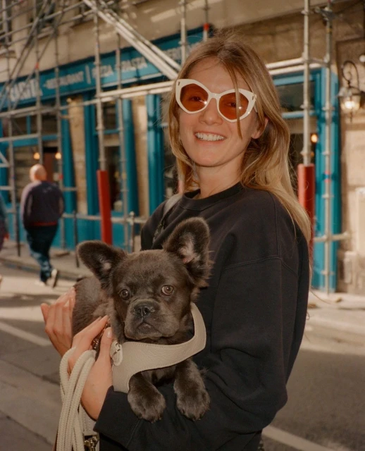 A woman in sunglasses smiles while holding a French Bulldog in front of a building with scaffolding and blue signage.