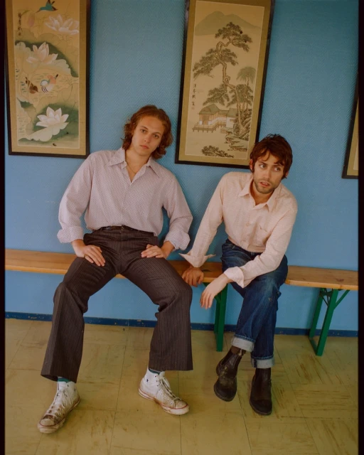 Two men sit on a bench in front of framed artwork. One man wears white sneakers; the other wears black boots. Light blue wall.
