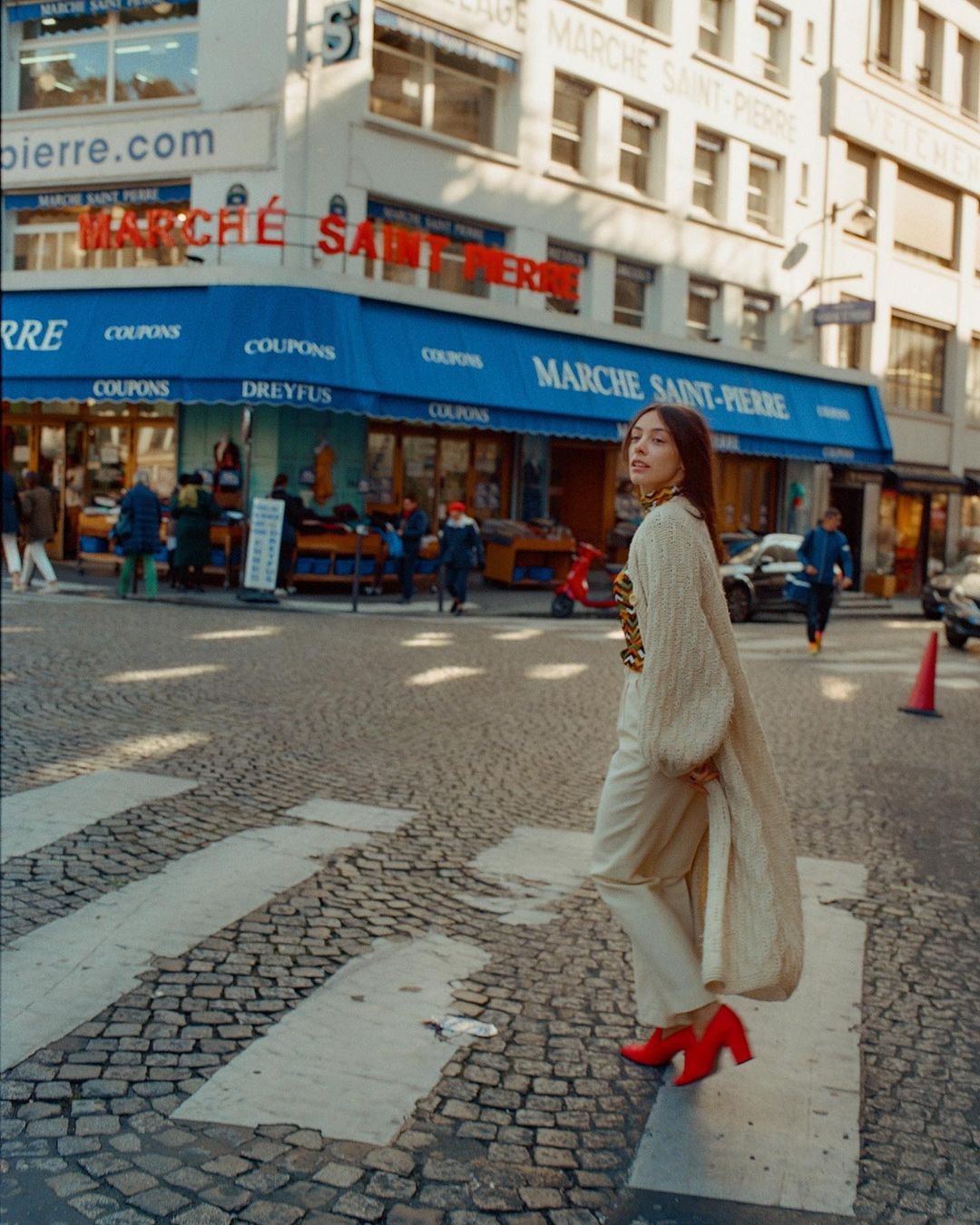 A woman walks across a zebra crossing in an urban setting, wearing a long coat and bright red heels, with buildings visible in the background.