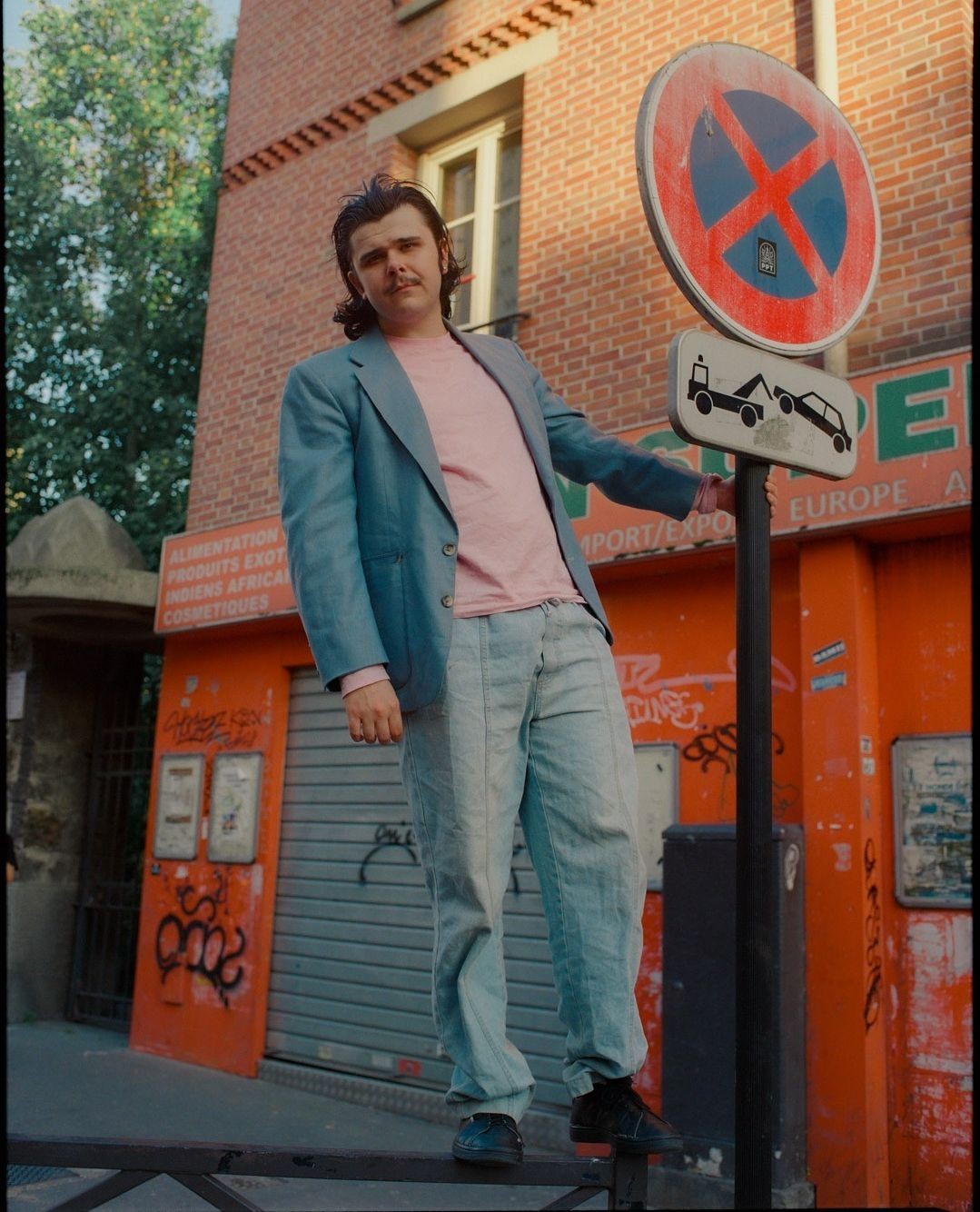 Adult male in a blue blazer and jeans poses next to a no parking sign in an urban setting with a brick building behind him.