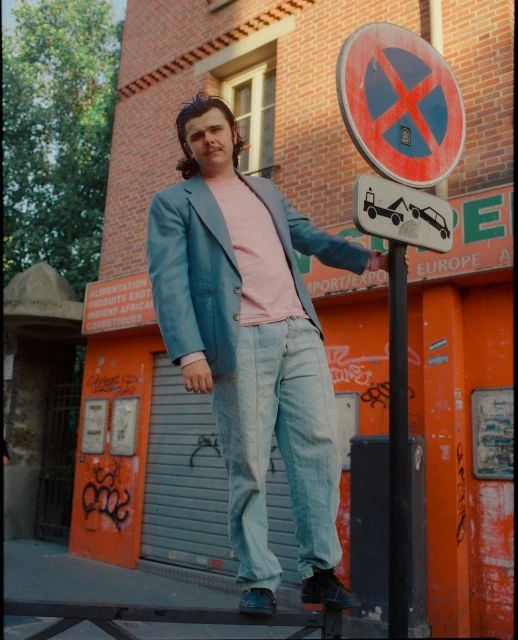 Adult male in a blue blazer and jeans poses next to a no parking sign in an urban setting with a brick building behind him.