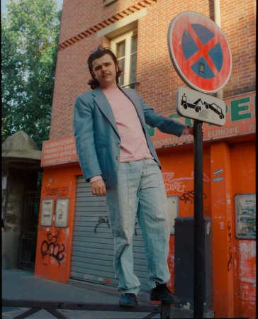 Adult male in a blue blazer and jeans poses next to a no parking sign in an urban setting with a brick building behind him.