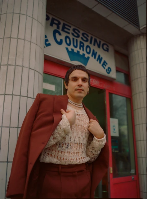 A man in a rust-colored suit and sheer sweater poses in front of a building with French signage. The photo is taken from a low angle.