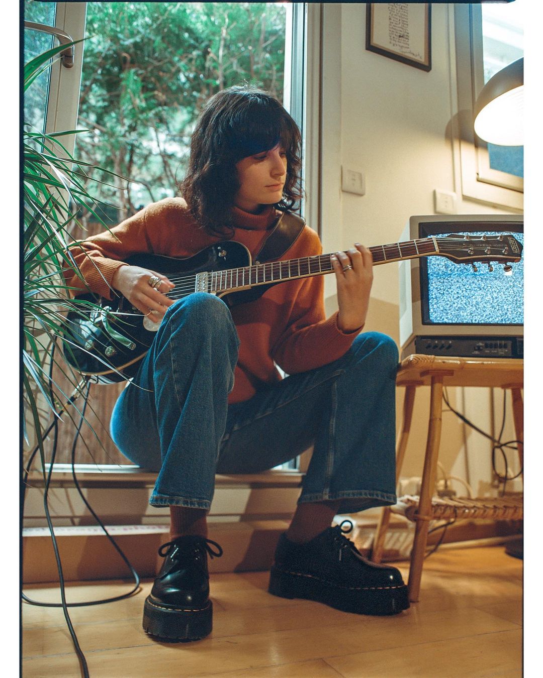 A young adult female sits indoors near a window and plays an electric guitar. A vintage television is in the background.
