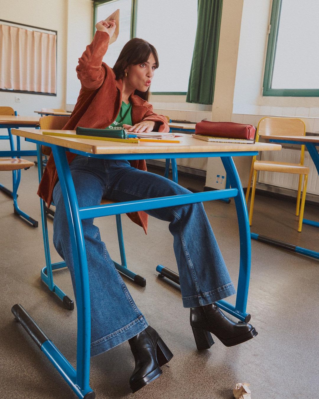 Young woman in classroom throwing crumpled paper at school desk, wearing brown jacket and jeans with black boots. Day.