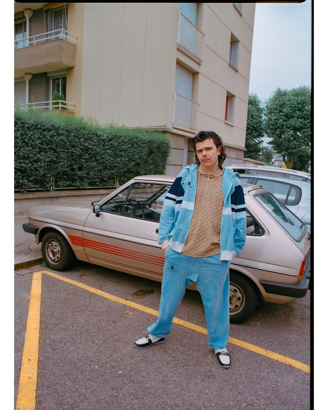 Man in tracksuit standing next to vintage car in urban parking lot. He wears a light blue tracksuit and black and white loafers.