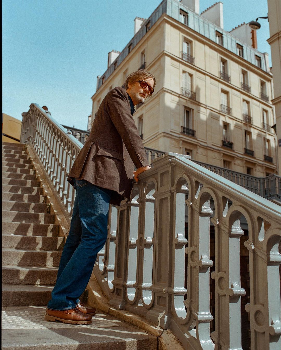 A man in a blazer standing on stairs with iron railings in an urban setting, framed by buildings and a clear blue sky on a sunny day.