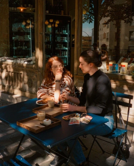 Two women sit at an outdoor cafe table with drinks and pastries, enjoying a sunny day with a building facade and tree branches visible.