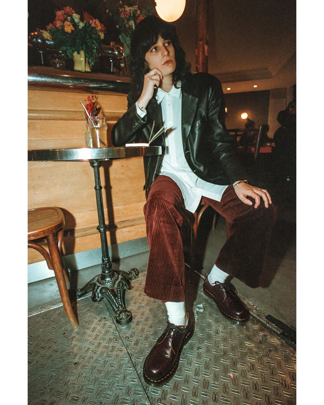 Adult male with curly hair sits at a metal table in an indoor environment wearing a black jacket, white shirt, and corduroy pants.