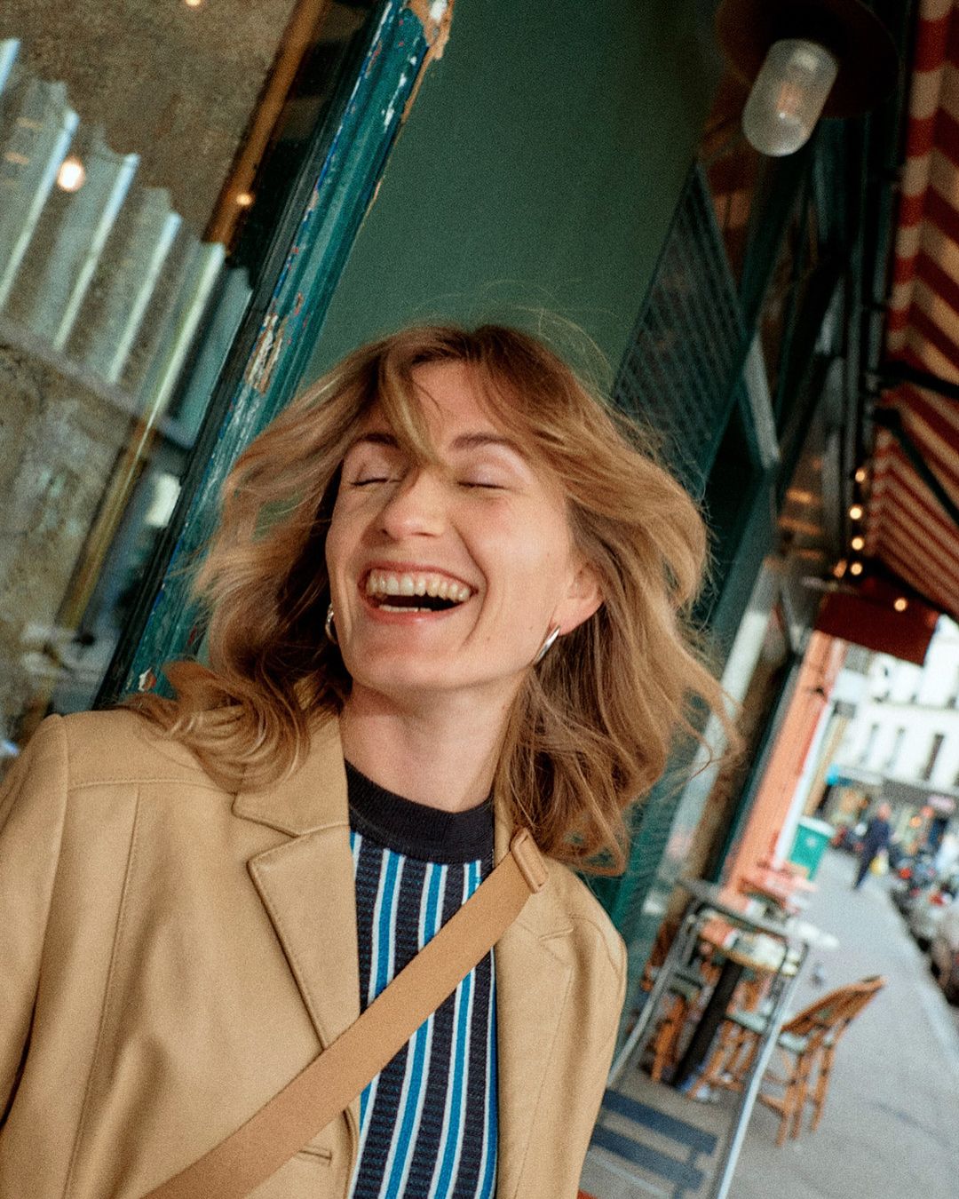 Smiling woman in a tan blazer walks along a European city street with outdoor cafe tables on the sidewalk and buildings in background.