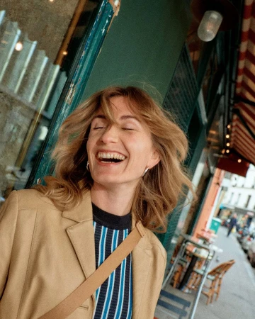 Smiling woman in a tan blazer walks along a European city street with outdoor cafe tables on the sidewalk and buildings in background.