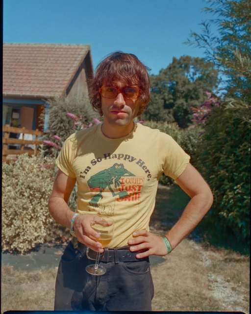 Man with tinted sunglasses holds drink outdoors. He wears graphic tee in garden setting with house and blue sky.
