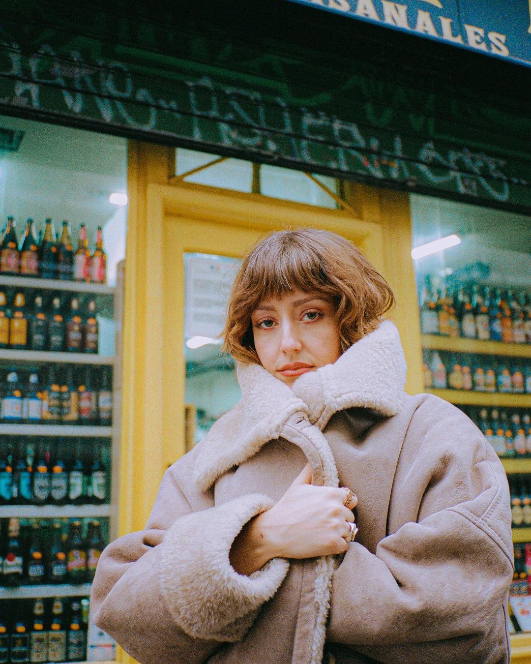 Woman in a beige coat stands in front of a shop with shelves of bottled drinks, creating a warm and inviting scene in an urban setting.