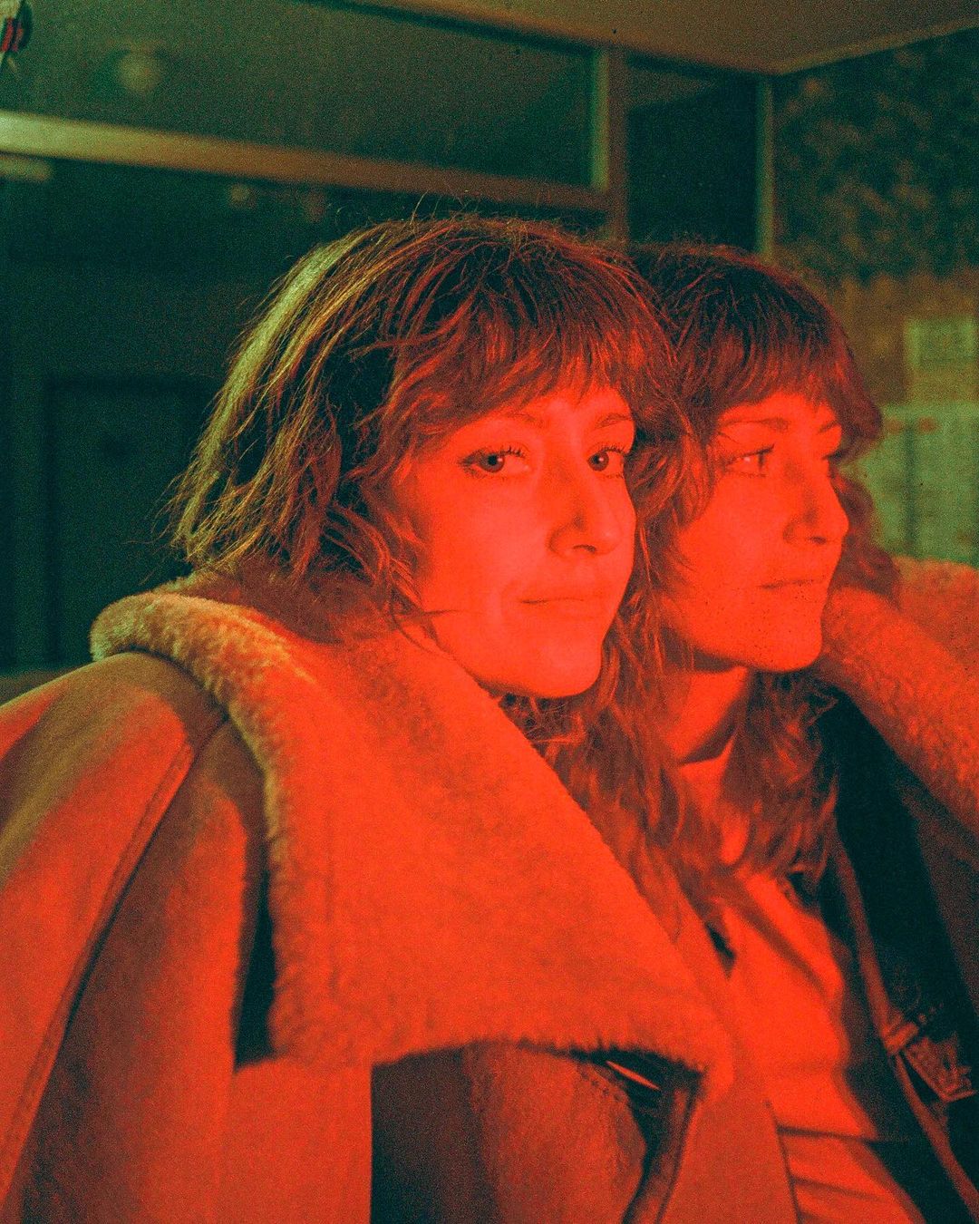 Portrait of a woman with auburn hair and a tan shearling coat, duplicated as if reflected in a mirror, under red and green lighting.