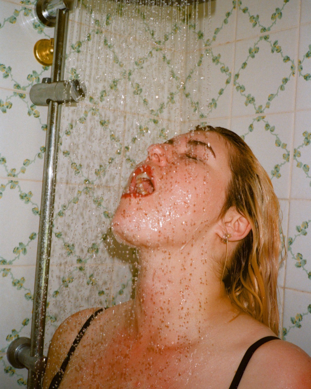 Woman with blonde hair stands under a shower with tiled walls, water cascading over her face, eyes closed, mouth open.