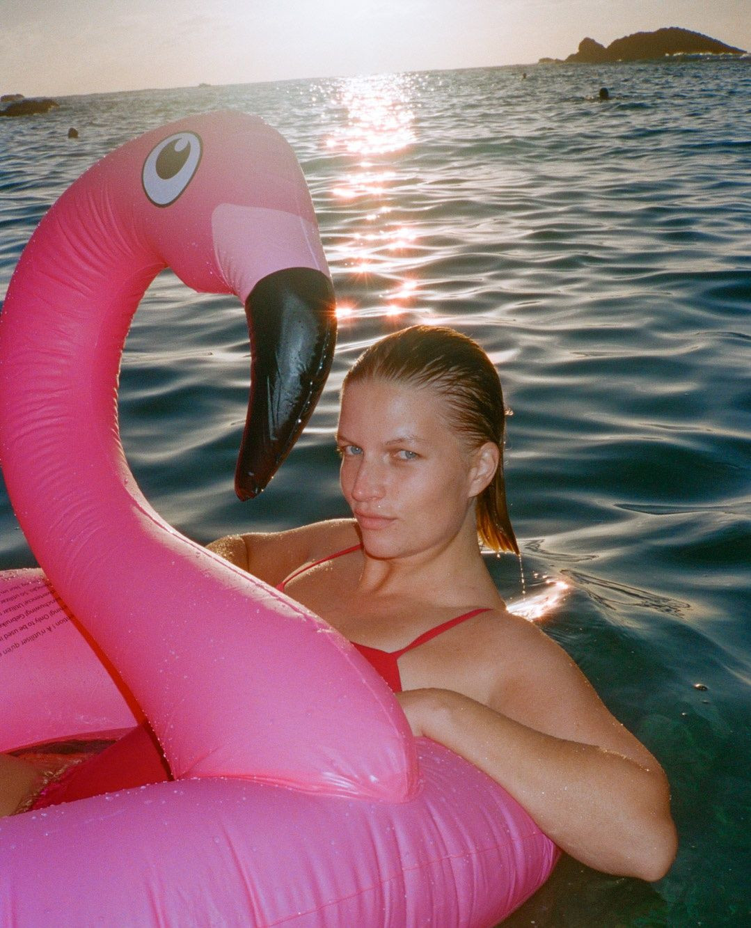 Blonde woman in a red swimsuit floats in the ocean with a large pink flamingo inflatable, enjoying a sunny day at the beach.