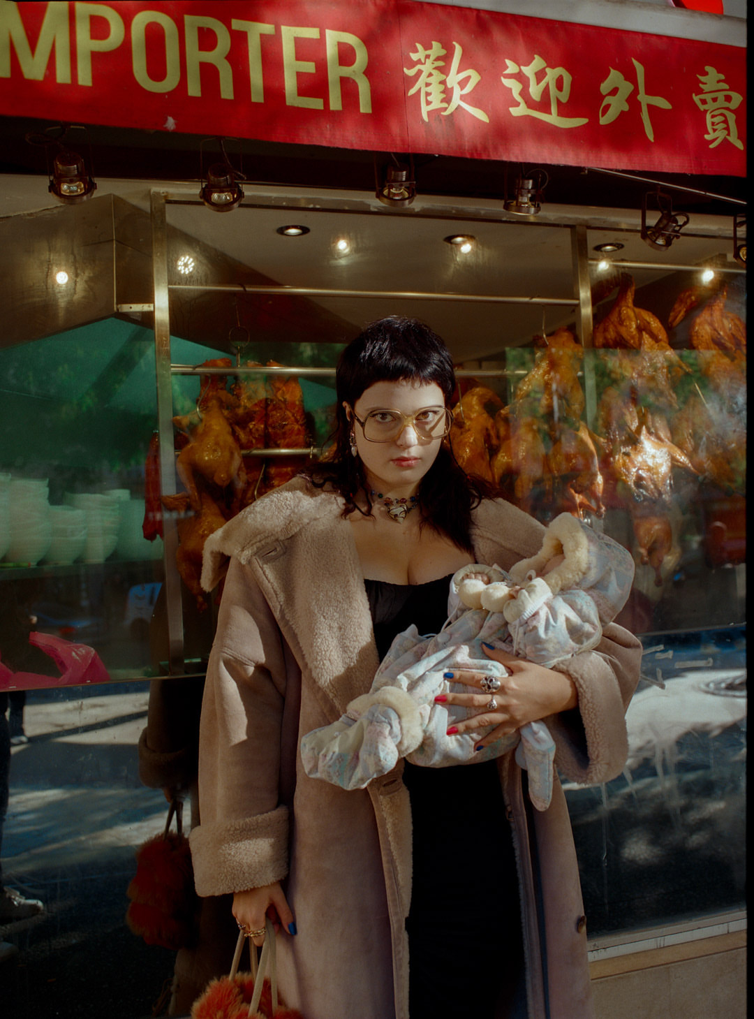 Woman in coat holds swaddled baby in front of restaurant with roasted fowl displayed in the window and Chinese signage overhead.