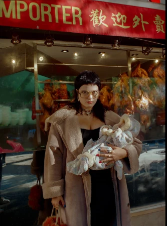 Woman in coat holds swaddled baby in front of restaurant with roasted fowl displayed in the window and Chinese signage overhead.