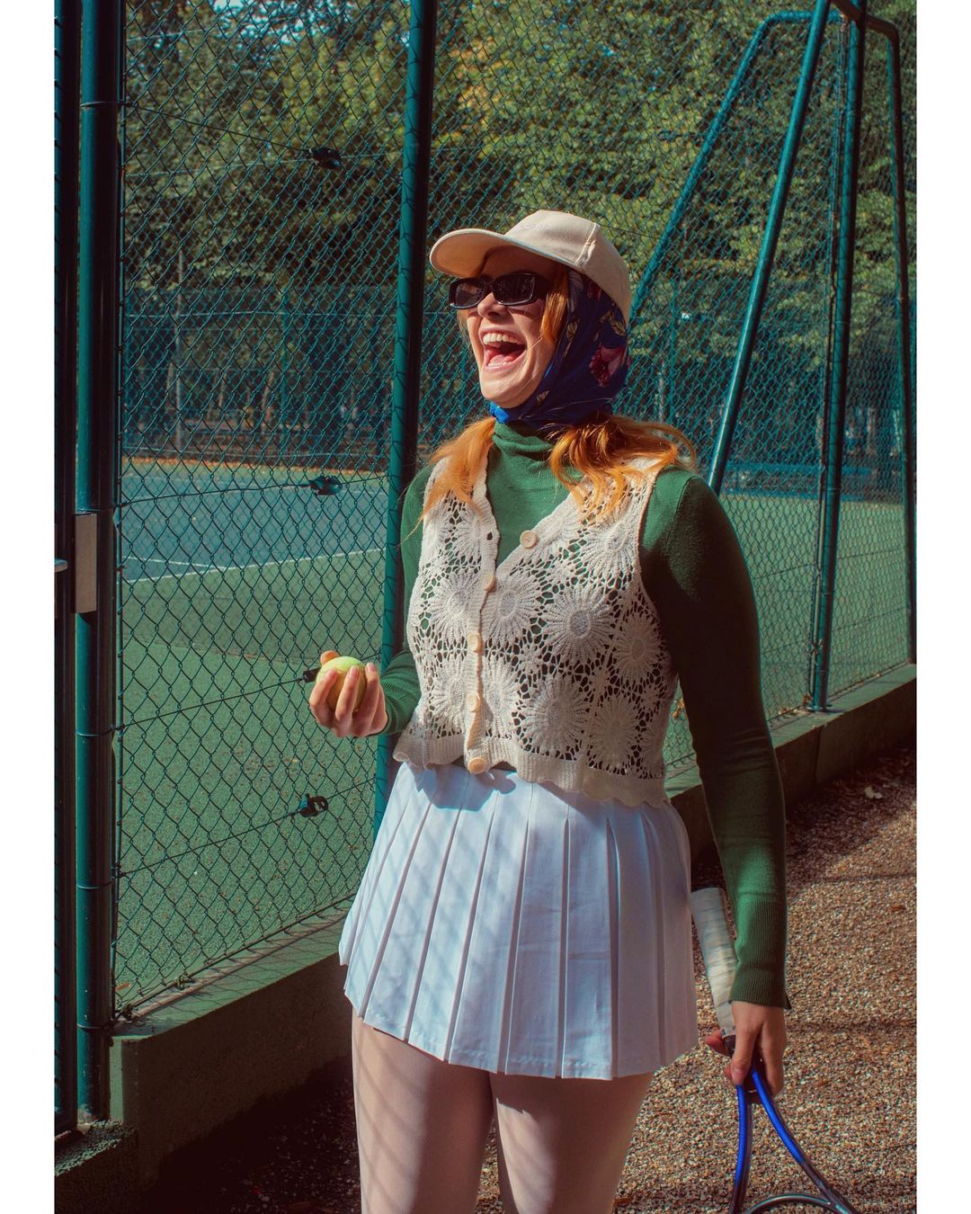 A woman smiles on a tennis court, wearing a white pleated skirt, a crochet vest, a baseball cap, and sunglasses, holding a tennis racket.