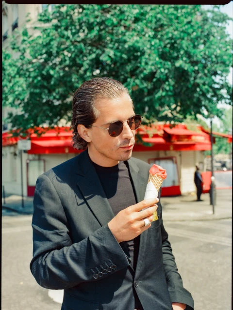 Adult male in a suit stands outdoors eating ice cream with green trees and a red building in the background on a sunny day.