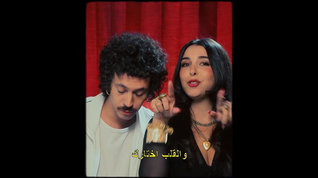 Music video scene with a young adult couple in a studio, red curtain backdrop, vintage filter, and Arabic text overlay at the bottom.