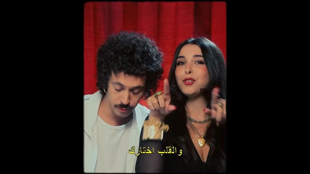 Music video scene with a young adult couple in a studio, red curtain backdrop, vintage filter, and Arabic text overlay at the bottom.