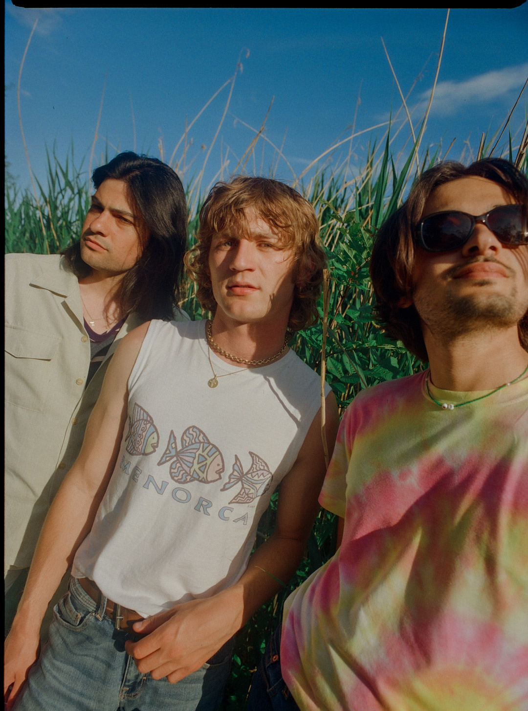 Three young adults pose outdoors in grass field under blue sky. The person in the center wears a white tank top.