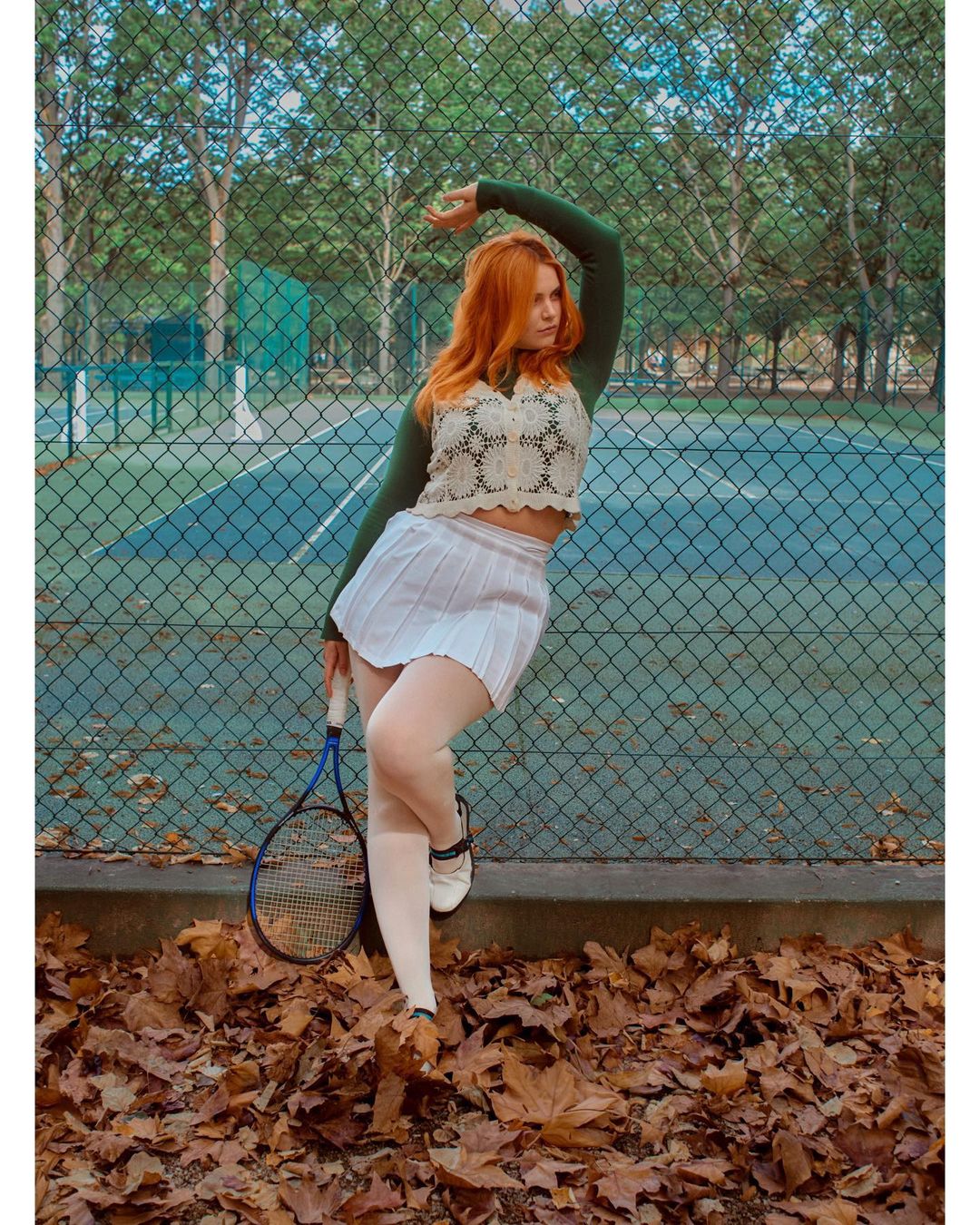 Woman poses next to tennis court wearing tennis skirt, top, and tennis shoes. She holds a tennis racket. Fallen leaves on the ground.