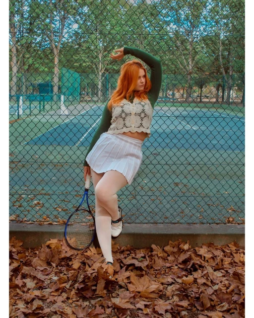 Woman poses next to tennis court wearing tennis skirt, top, and tennis shoes. She holds a tennis racket. Fallen leaves on the ground.