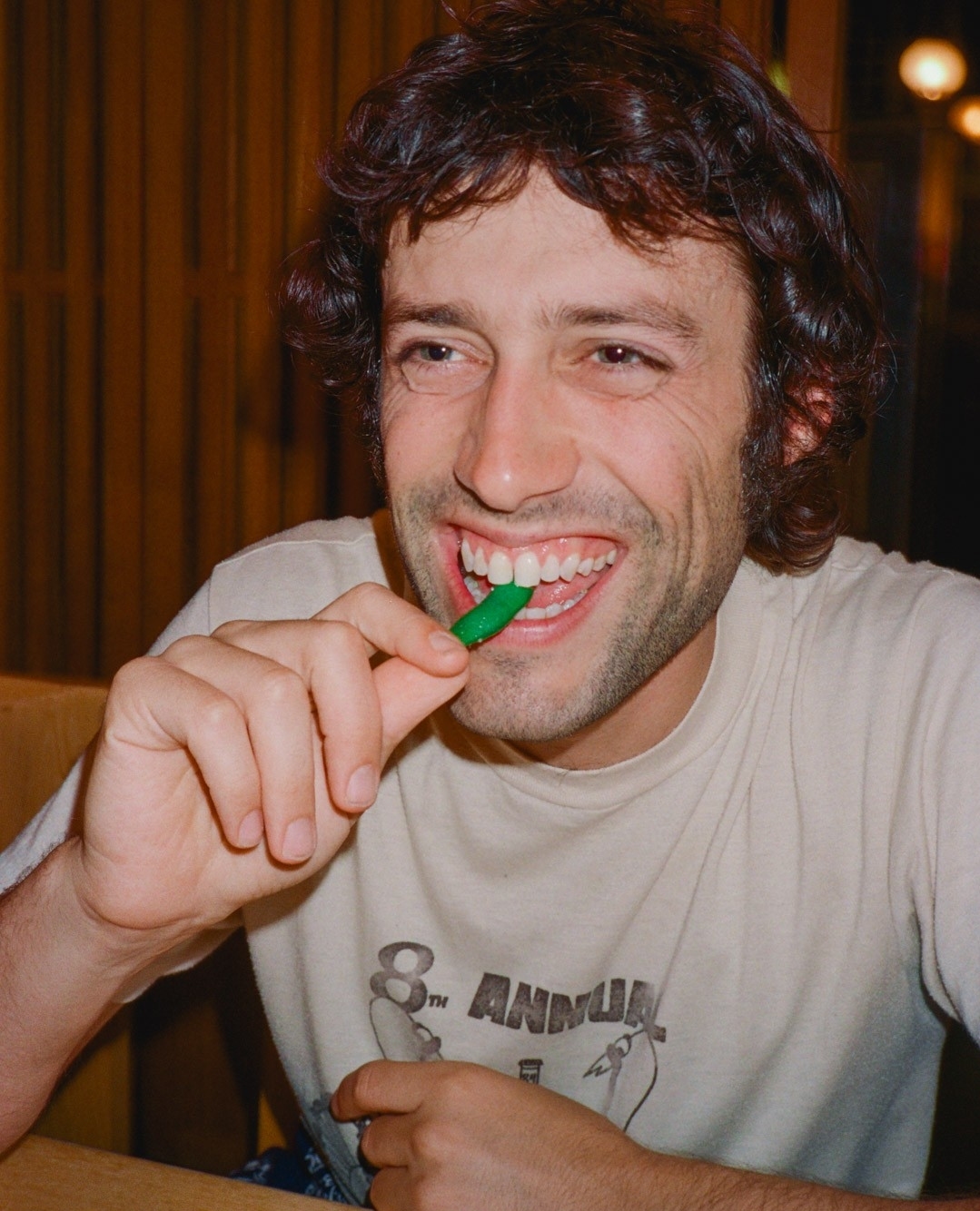 Dark-haired adult male with a slight beard smiles while eating a green candy stick; a portrait view.