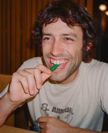 Man with dark curly hair wearing a white graphic t-shirt biting a green pepper while smiling at the camera