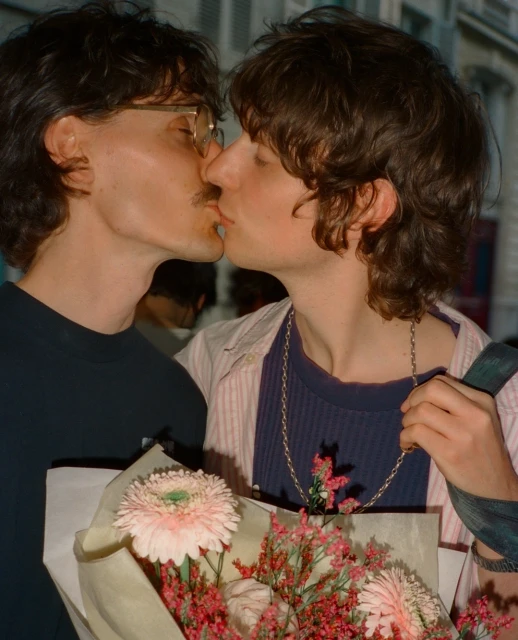 Close-up of two adult men kissing outdoors, one wearing glasses, with a bouquet of pink and white flowers in the foreground.