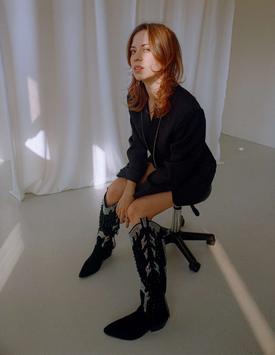 A woman with shoulder-length auburn hair sits on a stool in front of a white sheet wearing black boots and blazer in a studio setting.