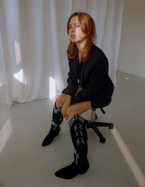 A woman with shoulder-length auburn hair sits on a stool in front of a white sheet wearing black boots and blazer in a studio setting.