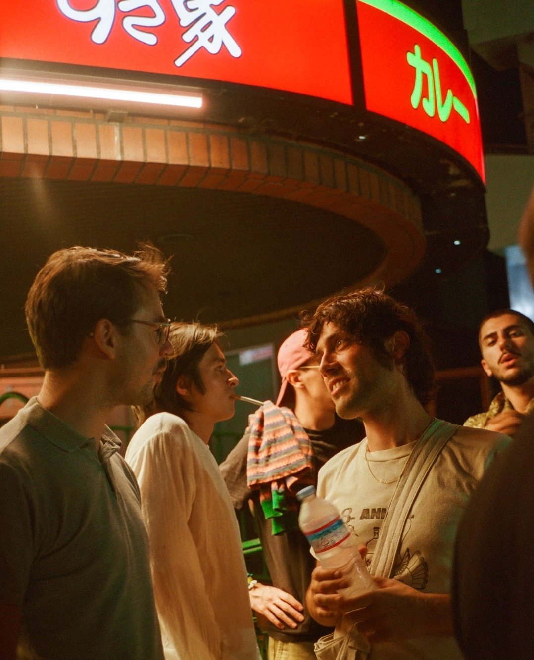 Group of young men in an outdoor urban setting, wearing casual clothing and facial expressions of conversation, in warm lighting.