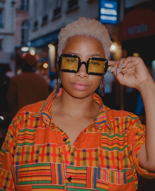 Portrait of a Black woman wearing orange patterned shirt, black glasses with yellow lenses, hoop earrings, looking at the viewer.