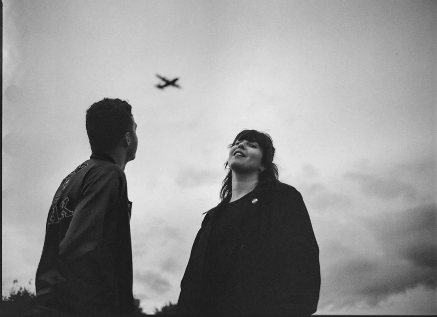 Two people look up at a plane in the sky against an overcast background in a black and white outdoor shot.