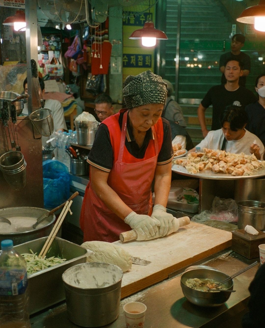 An Asian woman in an apron and headscarf uses a rolling pin at an indoor food stand, surrounded by bowls, utensils, and other people.