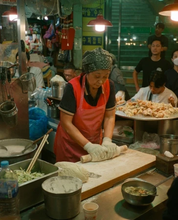An Asian woman in an apron and headscarf uses a rolling pin at an indoor food stand, surrounded by bowls, utensils, and other people.